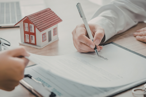 Pessoa a assinar um contrato junto de uma casa em miniatura sobre a secretária