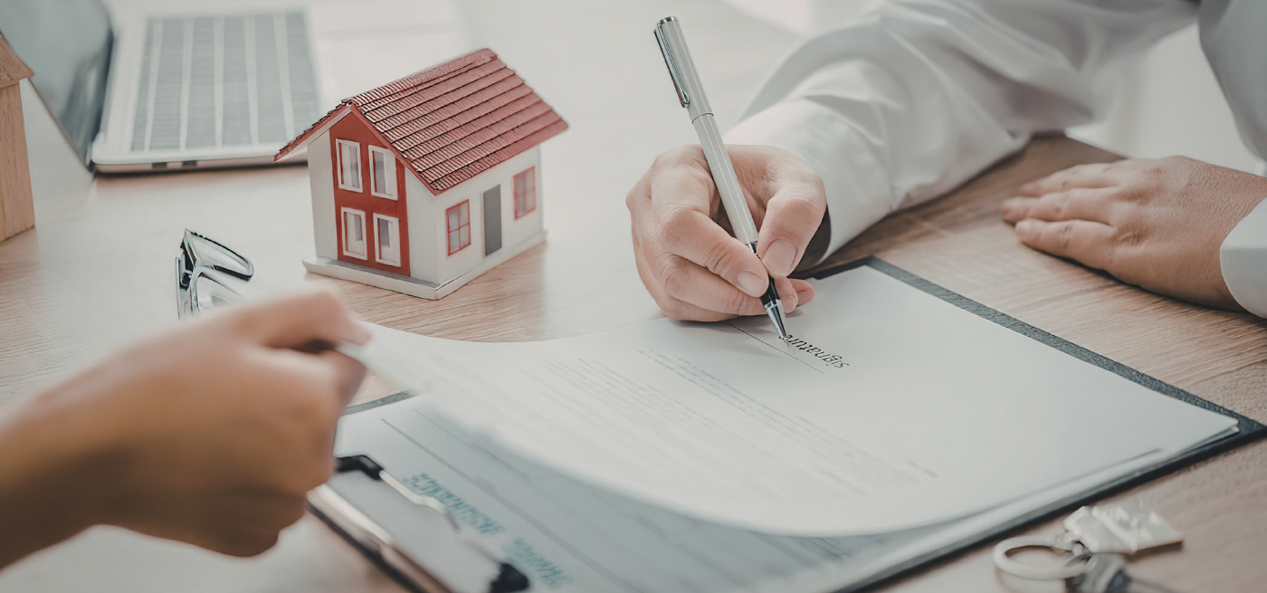 Pessoa a assinar um contrato junto de uma casa em miniatura sobre a secretária